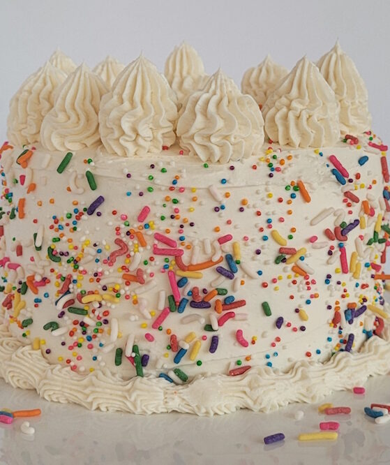 A vegan vanilla cake on a cake stand. The cake is covered in vegan sprinkles and has intricate piping on the top and base of the cake.