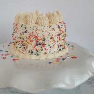 A vegan vanilla cake on a cake stand. The cake is covered in vegan sprinkles and has intricate piping on the top and base of the cake.