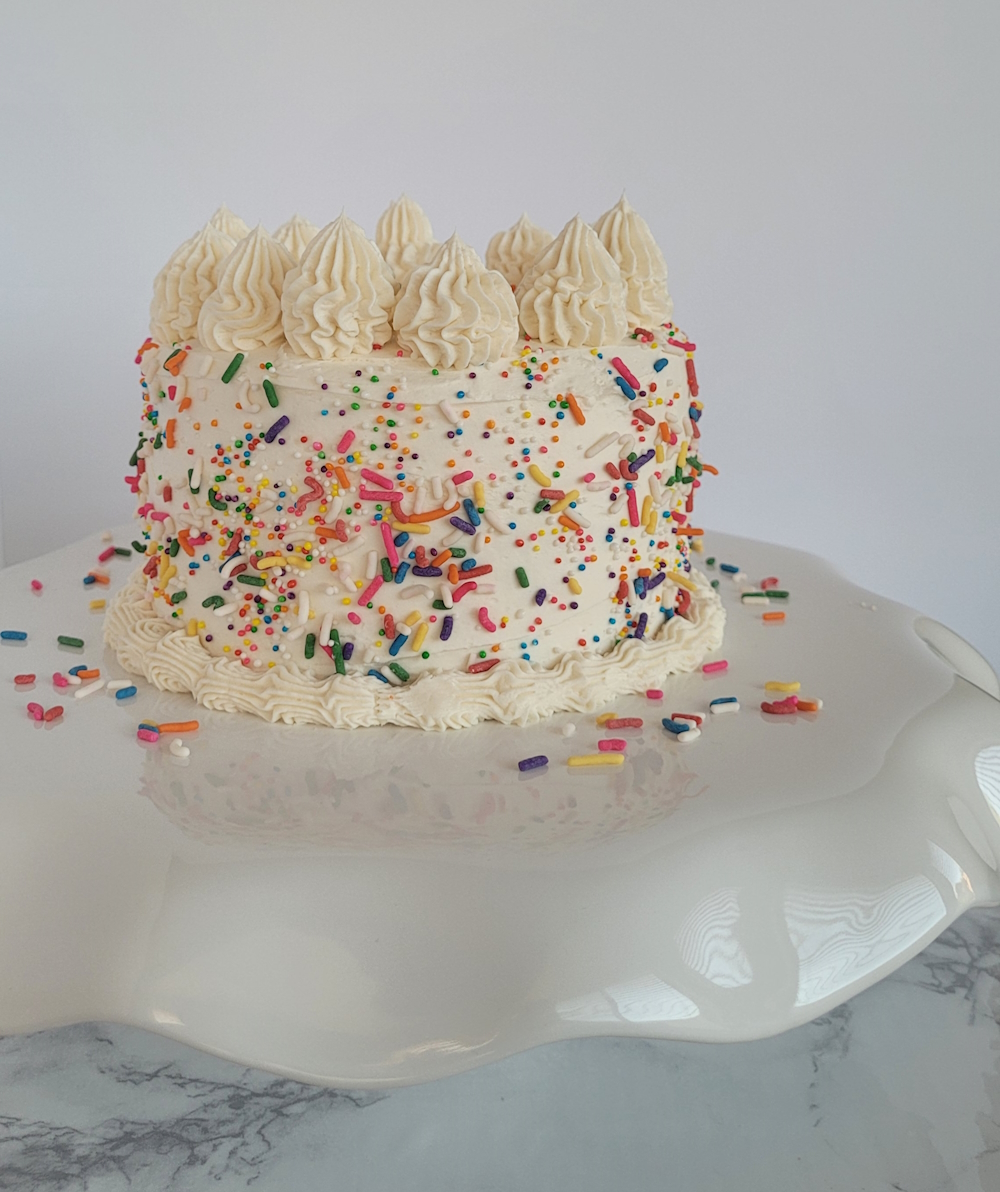 A vegan vanilla cake on a cake stand. The cake is covered in vegan sprinkles and has intricate piping on the top and base of the cake.